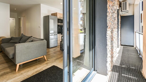 A modern living room with a sofa, a kitchen area, and a sliding glass door leading to a balcony.