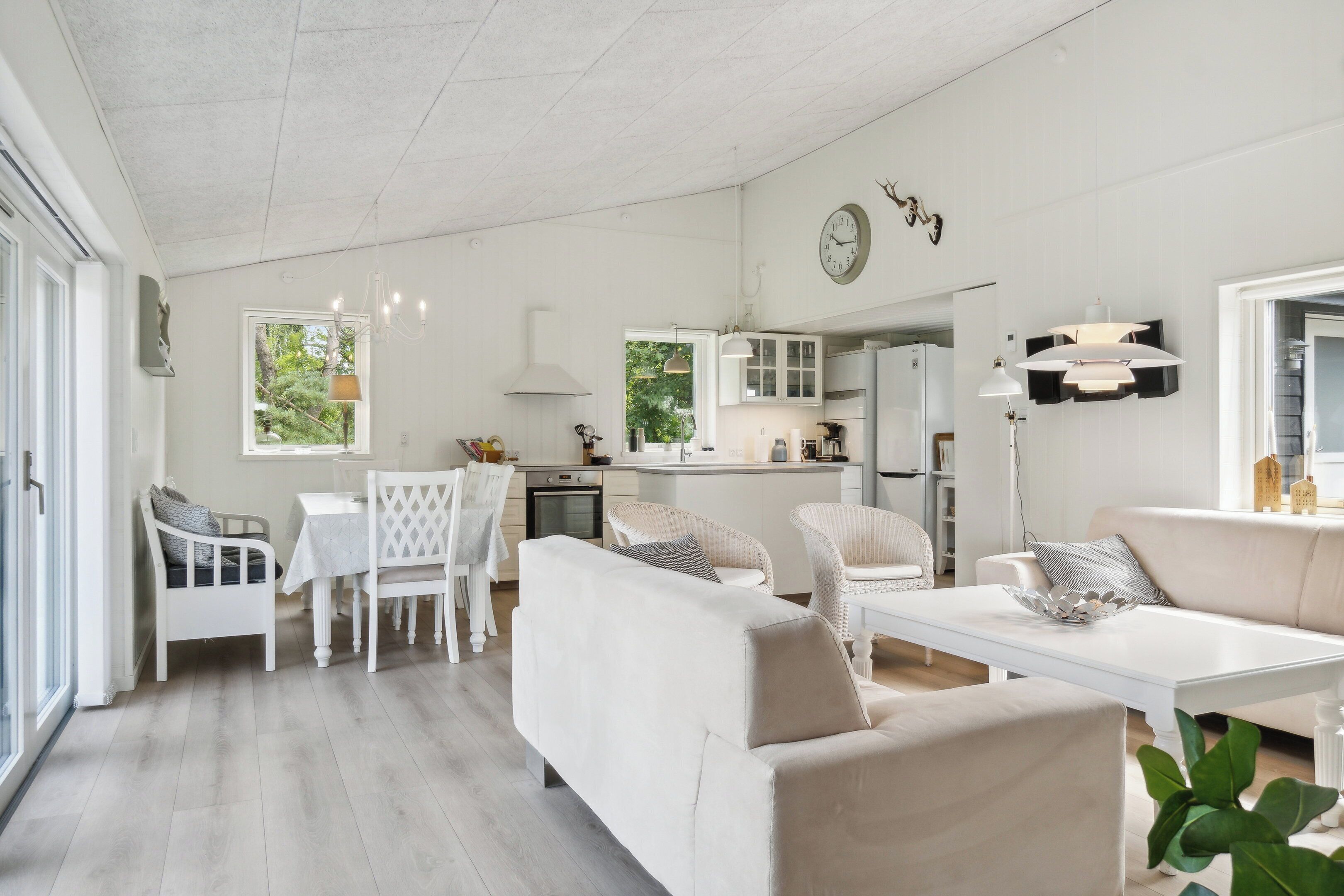 A spacious living area with a dining table, white furniture, and a kitchen in the background.