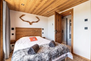 Un dormitorio con techo de madera, cama con manta blanca y gris, almohada con forma de ciervo y pared de piedra.