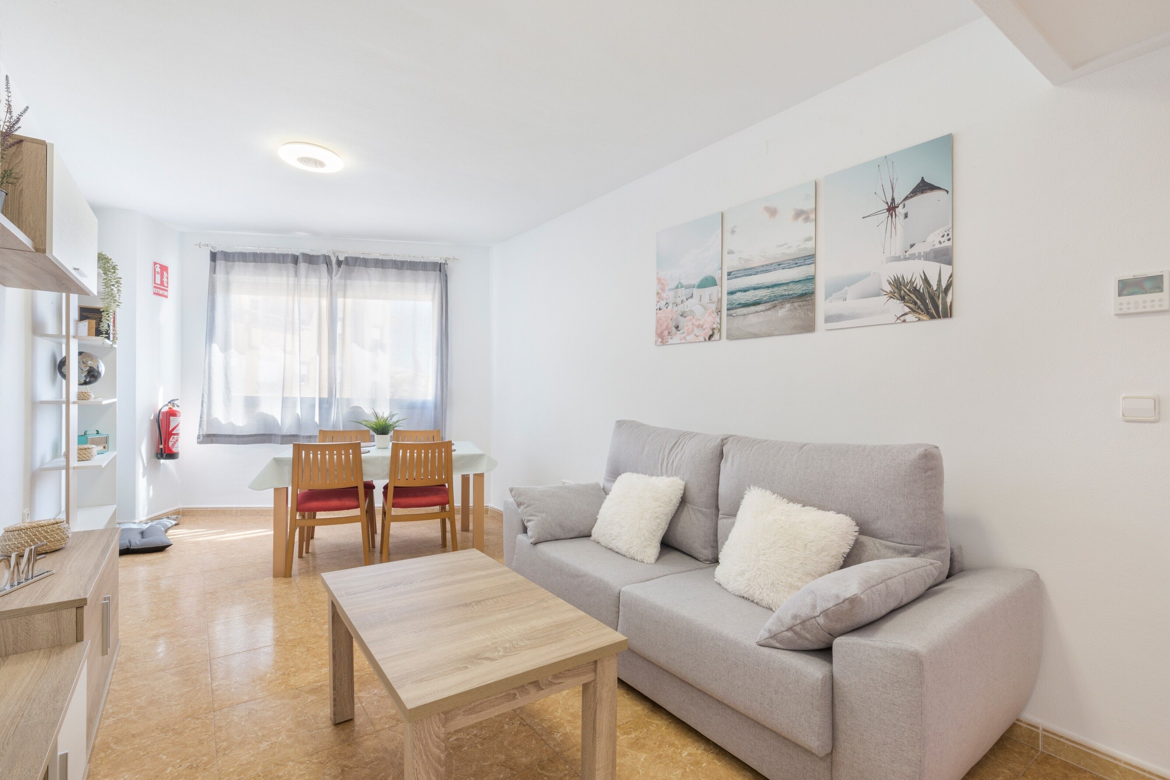 Una sala de estar con un sofá, una mesa de centro de madera, una mesa de comedor con sillas y un ventanal con cortinas traslúcidas.