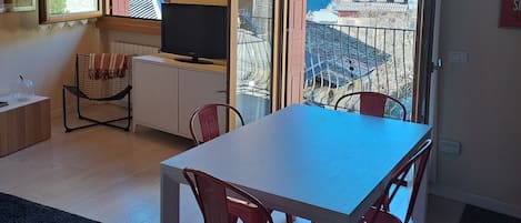 Un área de comedor con una mesa, sillas, un televisor y vistas a montañas a través de un ventanal.