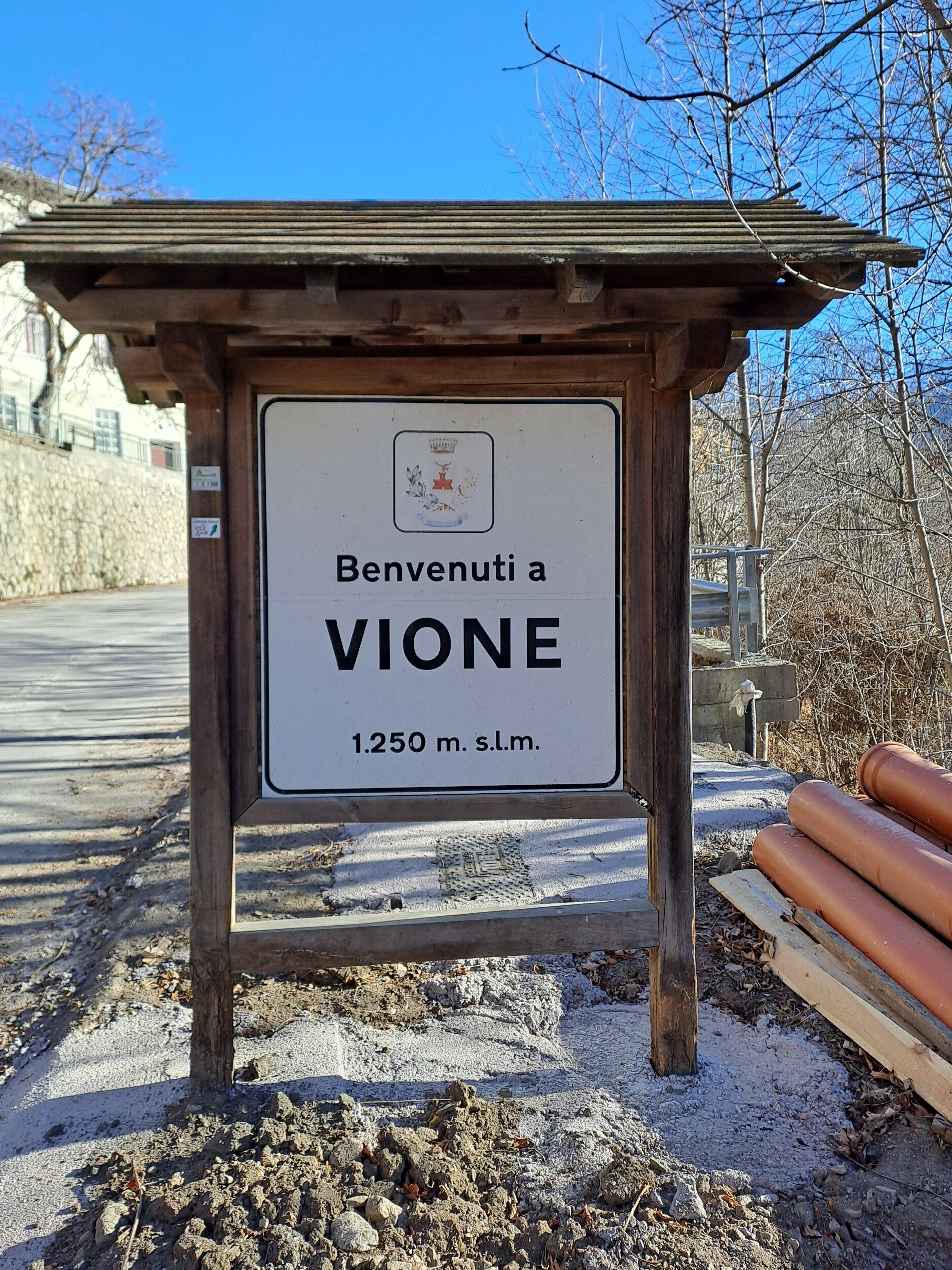 Willkommensschild für Vione, 1.250 Meter vom Aussichtspunkt entfernt, mit einer Steinmauer und kahlen Bäumen im Hintergrund.