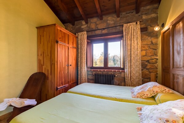 Una habitación con dos camas, armario de madera, pared de piedra, ventana con cortinas y un radiador.