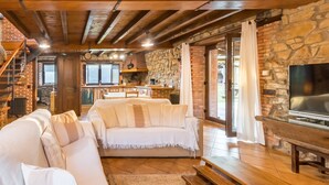 A cozy living room with a stone wall, wooden beams, a sofa, a TV, and a kitchen area. - Country House 'El Paraiso Del Yayo' with Mountain View, Private Terrace and Wi-Fi (Cantabria)