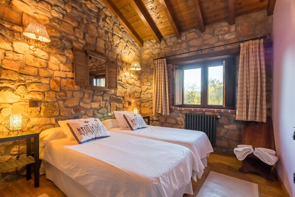 A room with stone walls, two beds with white bedding, a window with curtains, and a radiator. - Country House 'El Paraiso Del Yayo' with Mountain View, Private Terrace and Wi-Fi (Cantabria)