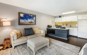 Een moderne woonkamer met een bank, voetenbankje en een keukenhoek met kasten en apparatuur.