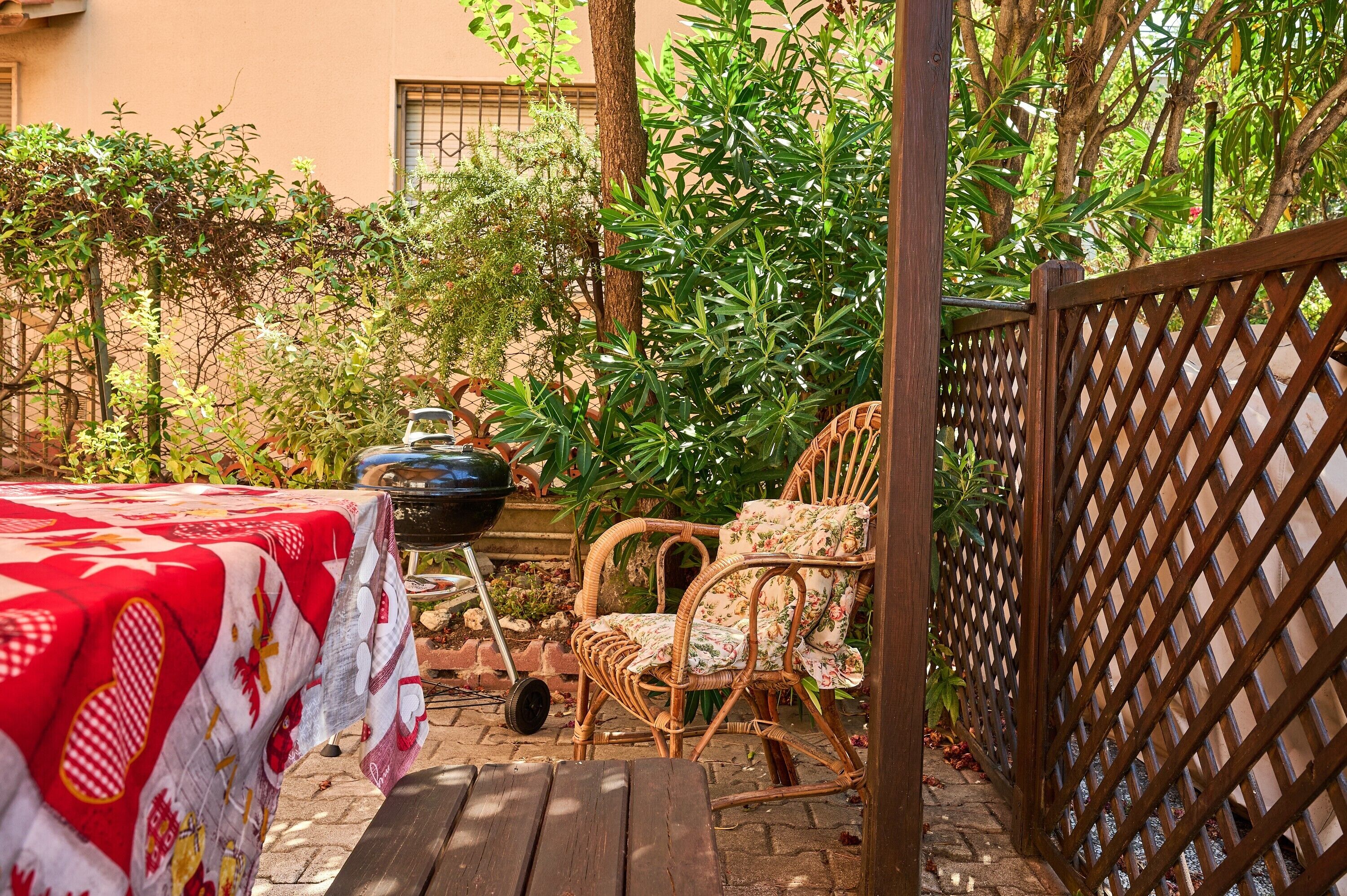 Une terrasse avec une table recouverte d’une nappe à carreaux rouges et blancs, une chaise en osier avec un revêtement à motifs floraux et un barbecue.