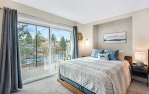 A bedroom with a large sliding glass door, a bed with patterned bedding, a nightstand with a lamp, and a framed picture on the wall.