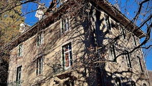 A historic stone building with multiple windows and a balcony, surrounded by trees and a garden.