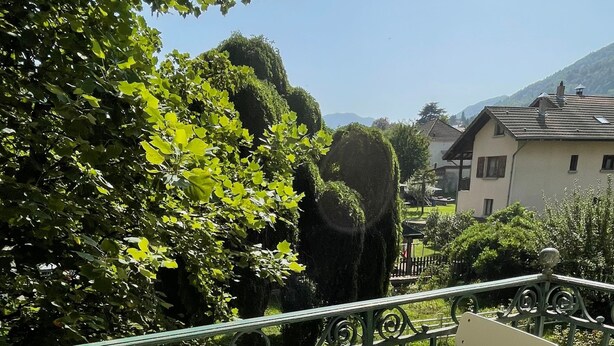 A balcony with a round table and two chairs, surrounded by greenery and a view of a house.