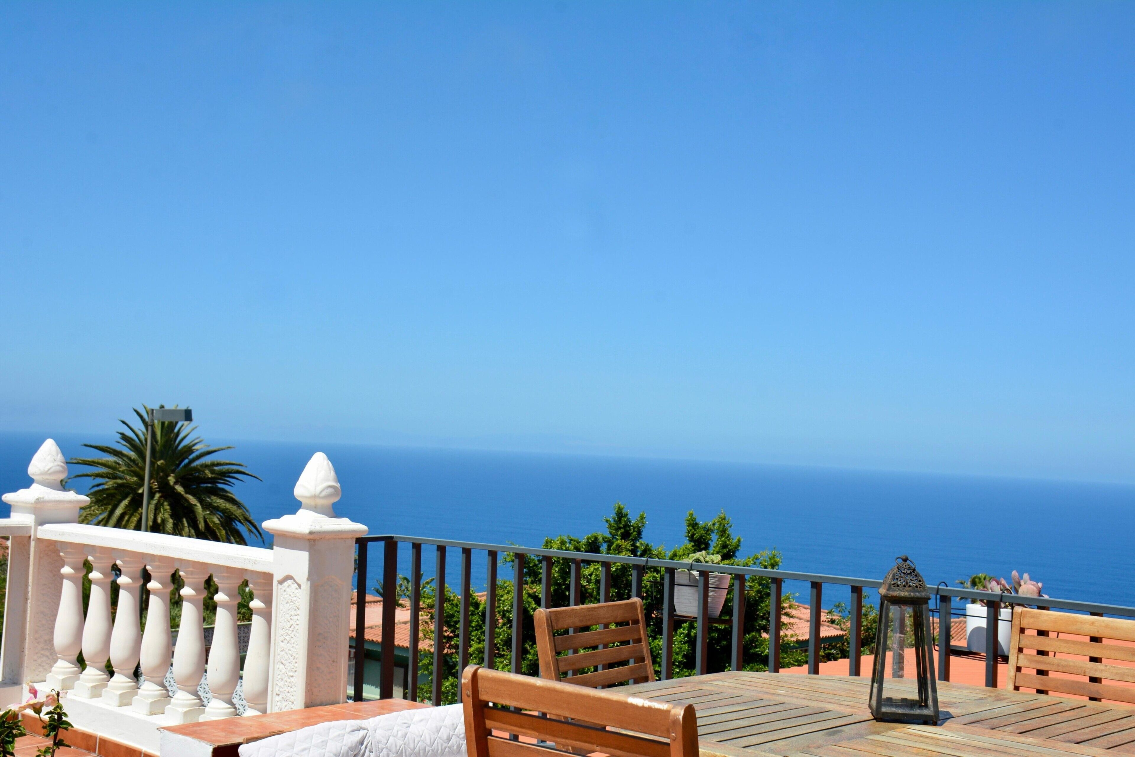 Uma varanda com mesa e cadeiras de madeira, guarda-corpo branco e vista para o mar.