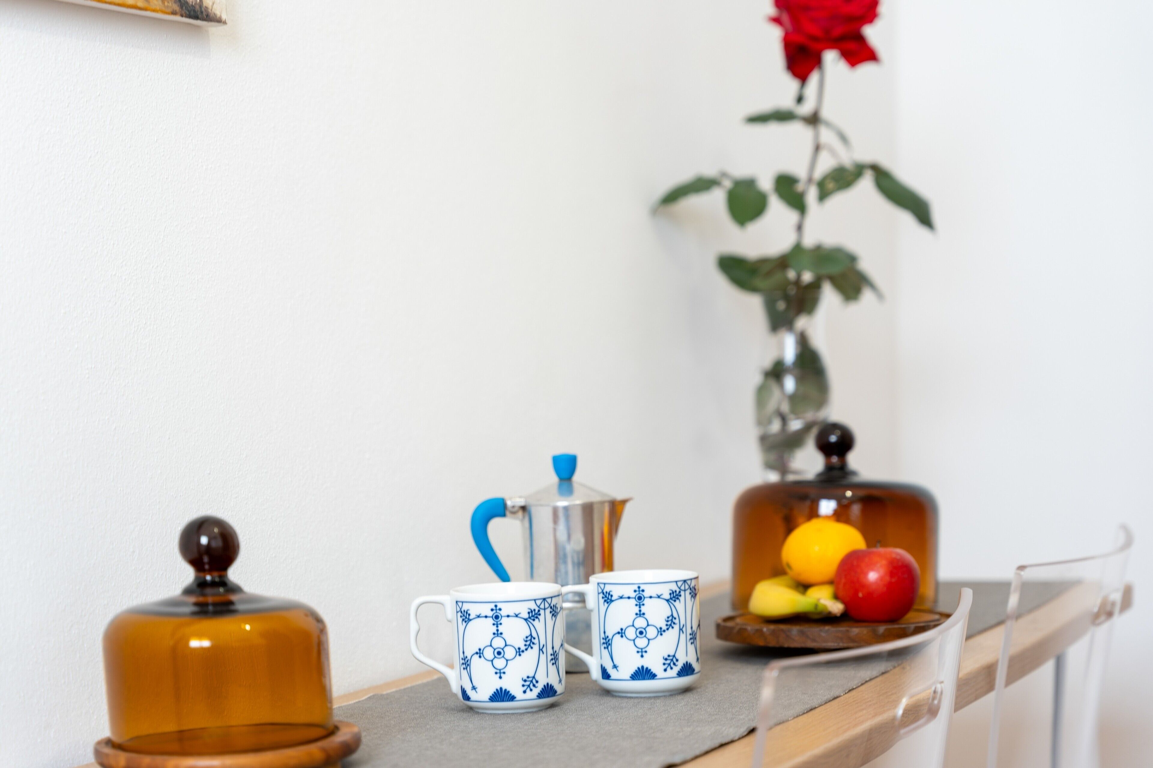 Una mesa con dos tazas decoradas, una cafetera, un recipiente de vidrio y un florero con una rosa roja.