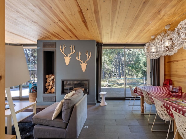 Ein modernes Interieur mit Holzbalkendecke, Kamin, einer Couch, einem Esstisch mit Stühlen und einem großen Fenster mit Blick ins Freie.