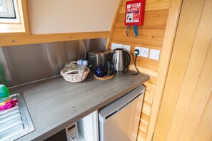 Ett litet kök med en brödrost, vattenkokare och kaffebryggare i rostfritt stål, placerade på en bänkskiva i trä. En brandfilt är upphängd på väggen ovanför bänkskivan.