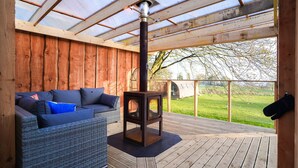 A wooden deck with a glass ceiling, a sofa with cushions, and a wood-burning stove.