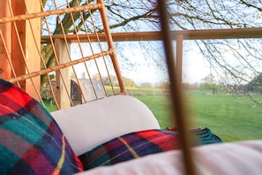 A wicker chair with a white cushion, a plaid blanket, and a view of a grassy field through a window.