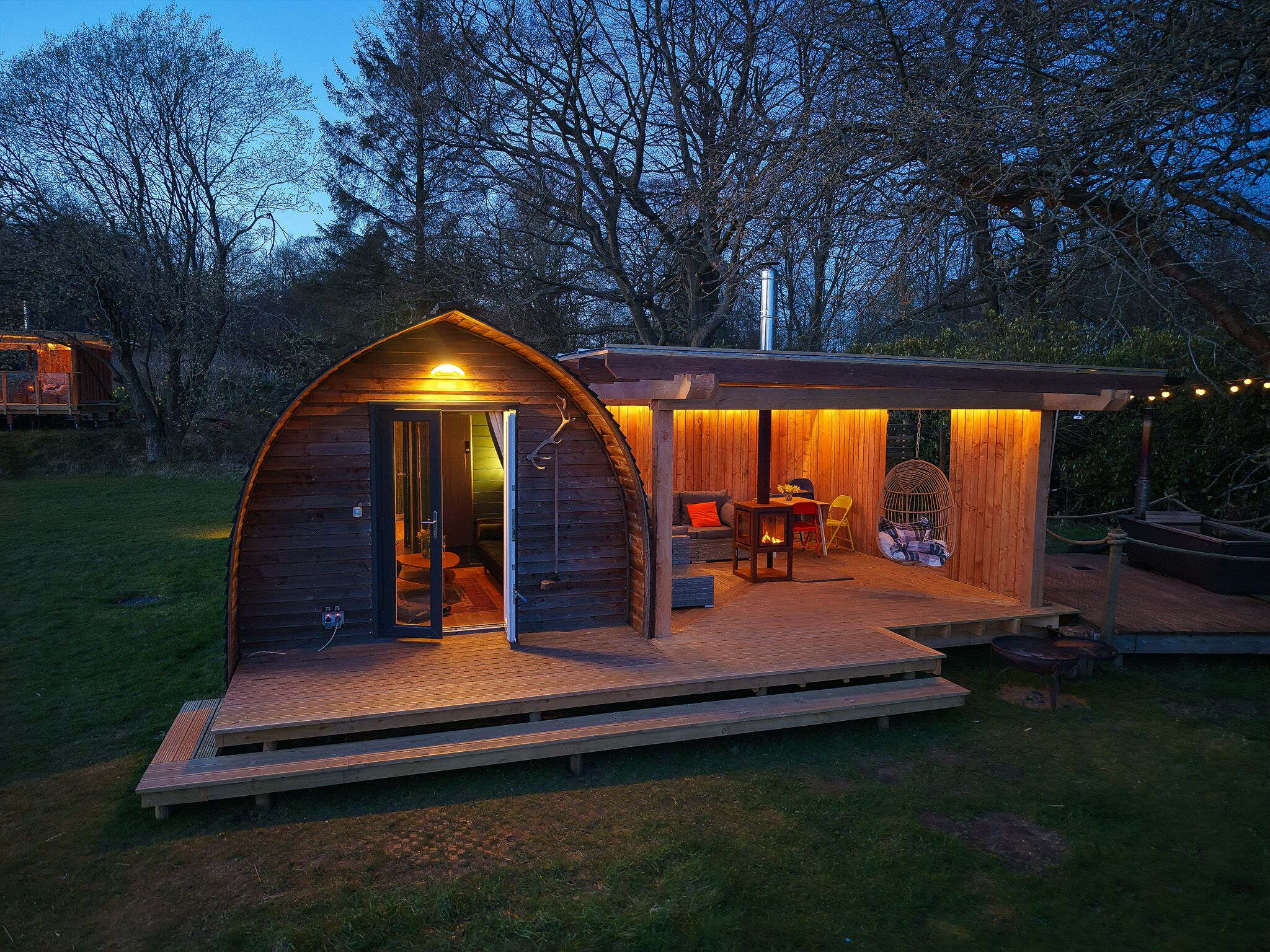 Een gezellig houten huisje met een boogvormige ingang, een klein terras met zitplaatsen en een vuurplaats.