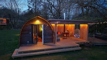 Een gezellig houten huisje met een boogvormige ingang, een klein terras met zitplaatsen en een vuurplaats.