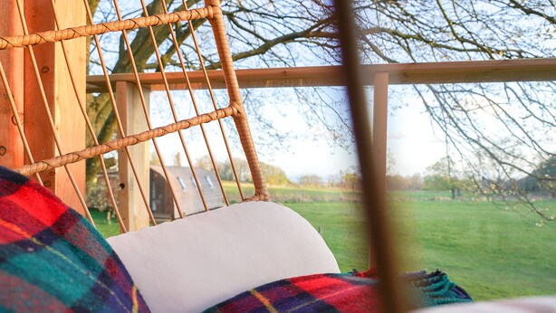 Una silla de mimbre con un cojín blanco, una manta a cuadros y vistas a un campo verde a través de una ventana.
