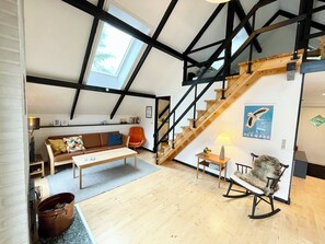 A loft-style room with exposed beams, a wooden staircase, a sofa, a rocking chair, and a poster on the wall. - Cozy, original holiday home - close to the beach (Væggerløse)
