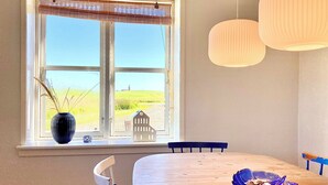 Un comedor con un gran ventanal, una mesa de madera, sillas, un jarrón con plantas y lámparas colgantes.