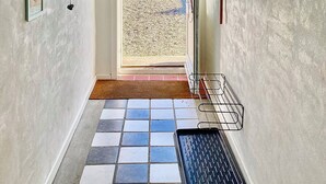 Un pasillo con una puerta que da al exterior, piso de baldosas, un felpudo negro, un perchero en la pared con objetos y cuadros enmarcados.