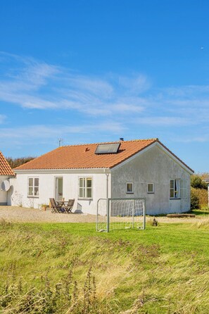 Une maison de plain-pied avec un toit recouvert de tuiles rouges, un petit patio avec une table et des chaises, et une porte bien visible.