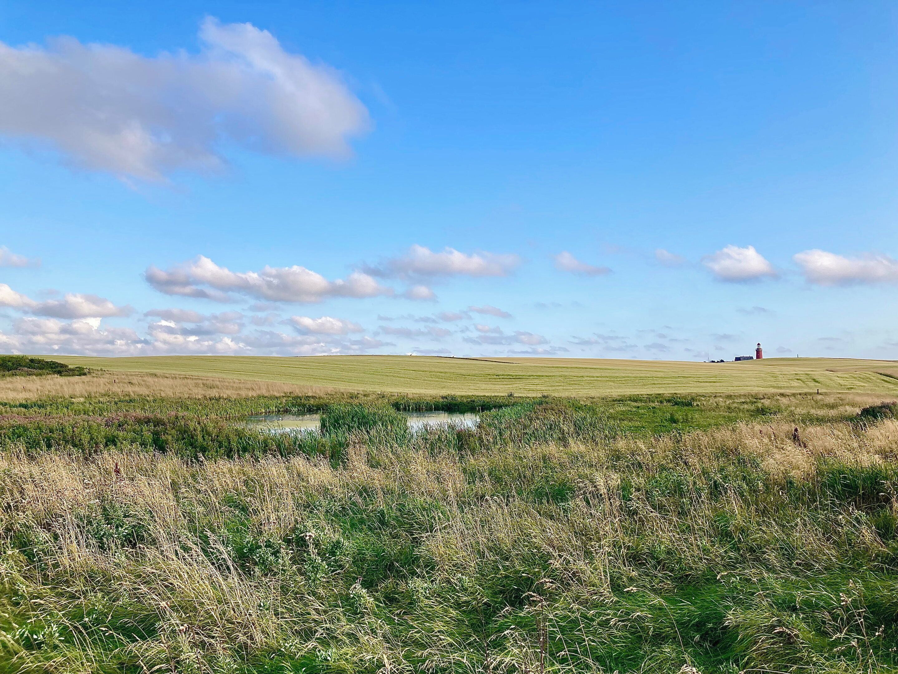 A flat landscape with a water body in the foreground, a distant red structure, and a clear sky with clouds.