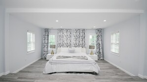 Ein Schlafzimmer mit einem großen Bett, weißen Wänden und Holzboden. Zwei Nachttische mit Lampen stehen bereit, und ein Fenster mit Jalousien sorgt für Licht.