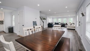 Ein geräumiger Essbereich mit einem langen Holztisch und weißen Stühlen, eine Küchenzeile mit weißen Schränken sowie ein Wohnzimmer mit einer Couch und einem Couchtisch.