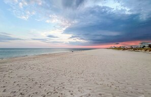 A wide sandy beach with footprints, a calm sea, and a cloudy sky.