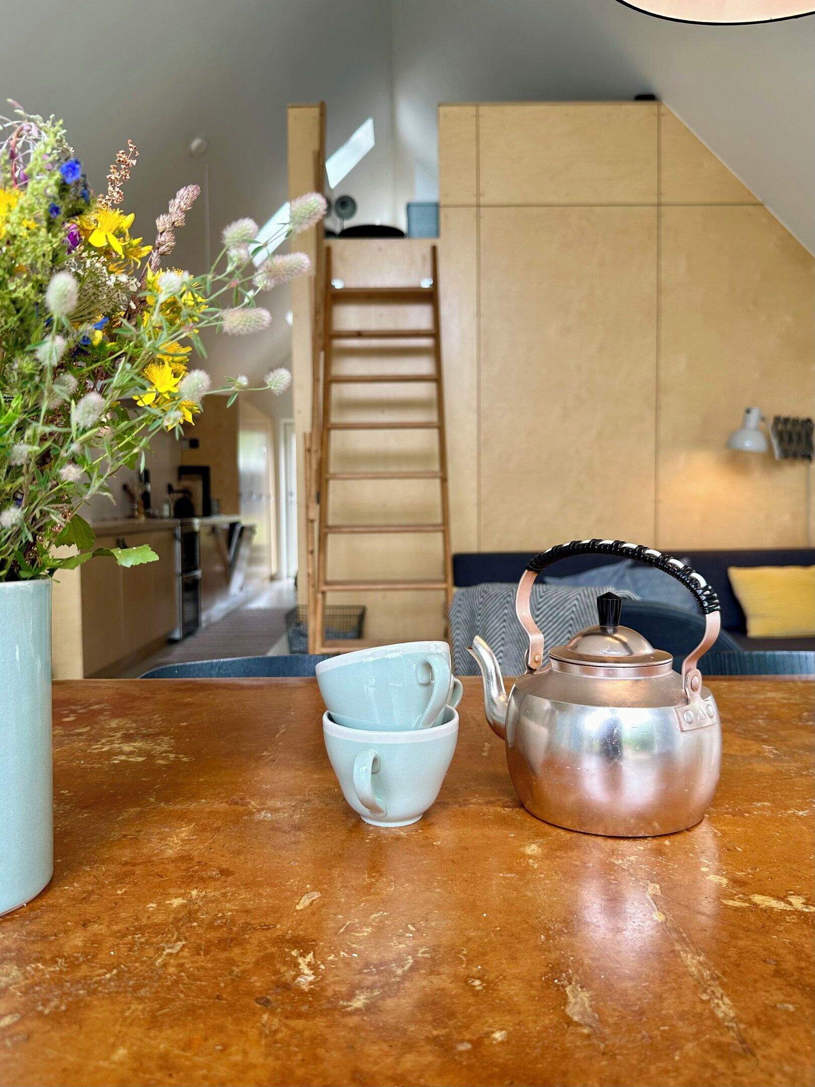 Eine Küche mit einer Holzleiter, einer weißen Vase mit Blumen und einem Wasserkocher auf einem Holztisch.