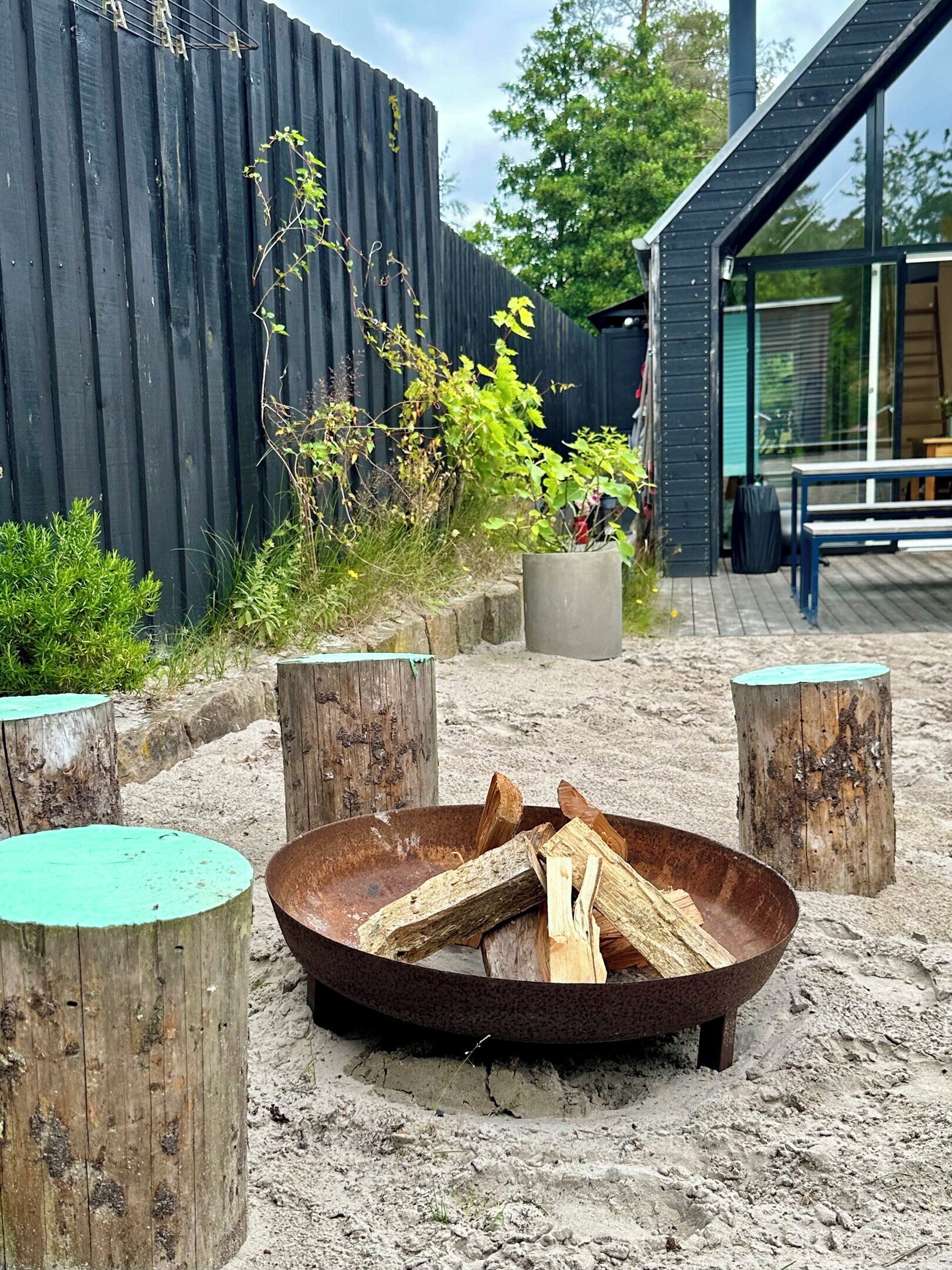 A fire pit with wood, surrounded by wooden stumps, in a sandy outdoor area with a wooden fence and a building with large windows in the background.