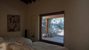 A bedroom with two beds, a wooden ceiling, a window with a view of trees and outdoor containers, and a wall with a colorful painting. - Zapote House (Malinalco)