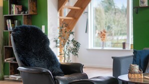 A room with a black leather chair, a wooden bookshelf, a window with a view of trees, and a table with a woven basket. - Modern house with space for relaxation (Aakirkeby)