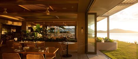 Un área de comedor al aire libre con mesas y sillas, vista al mar y un edificio de diseño moderno.