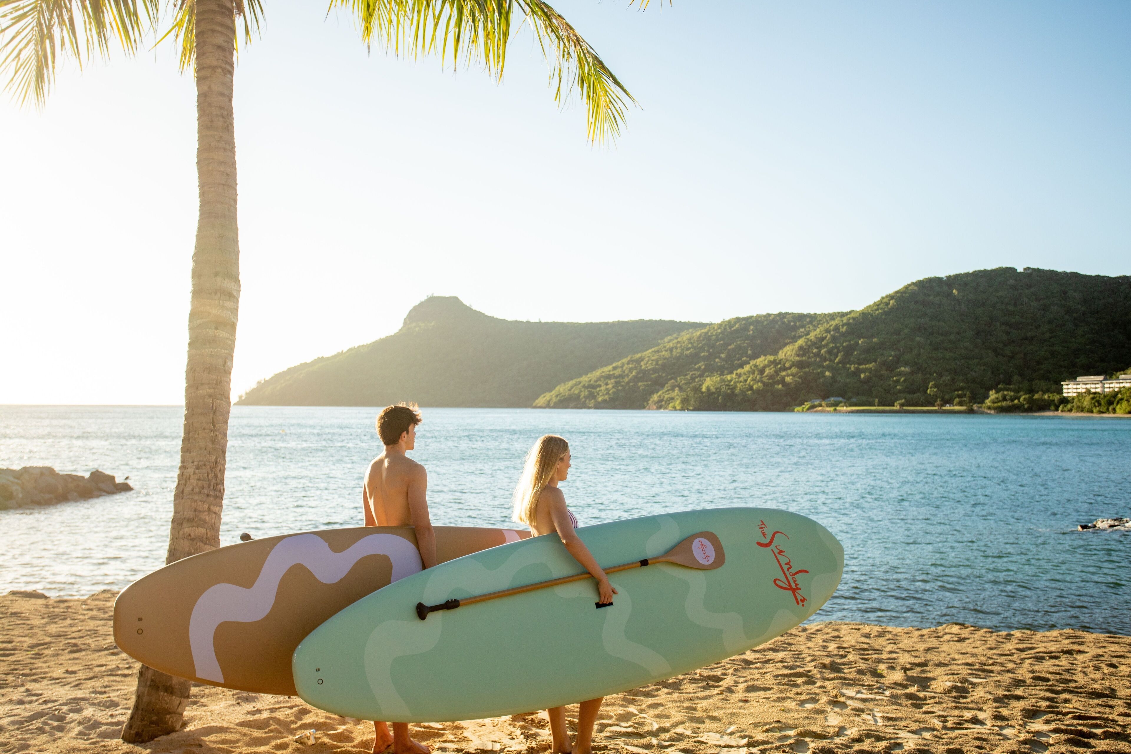 A beach scene with two people holding surfboards, palm trees, and a mountainous backdrop.