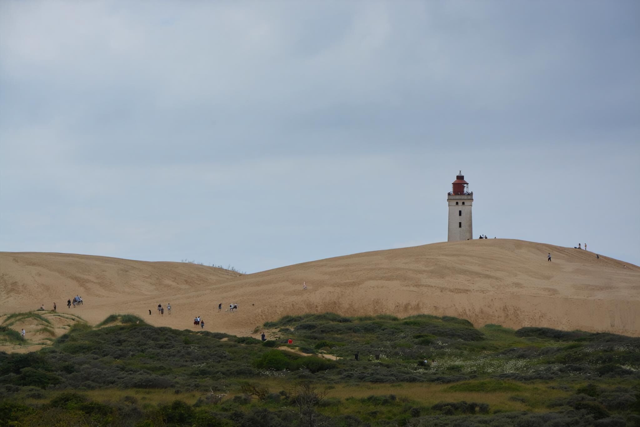 Un phare se dresse sur une colline de sable où la végétation est clairsemée, et il est entouré de quelques personnes.