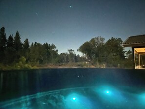 Une vue nocturne d’une piscine éclairée par deux projecteurs, entourée d’arbres et d’un bâtiment.