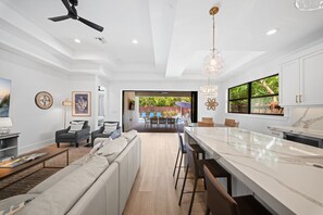 A modern kitchen with a large island, a dining area with a view of the outdoors, and a living room with a sofa and chairs. - LUXE: Home w/ 3 Kings, Pool & Spa, Bikes, BBQ (Naples)