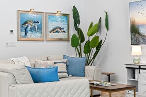 A living room with a white sofa, blue cushions, a coffee table, and two framed paintings of sea turtles. - LUXE: Home w/ 3 Kings, Pool & Spa, Bikes, BBQ (Naples)