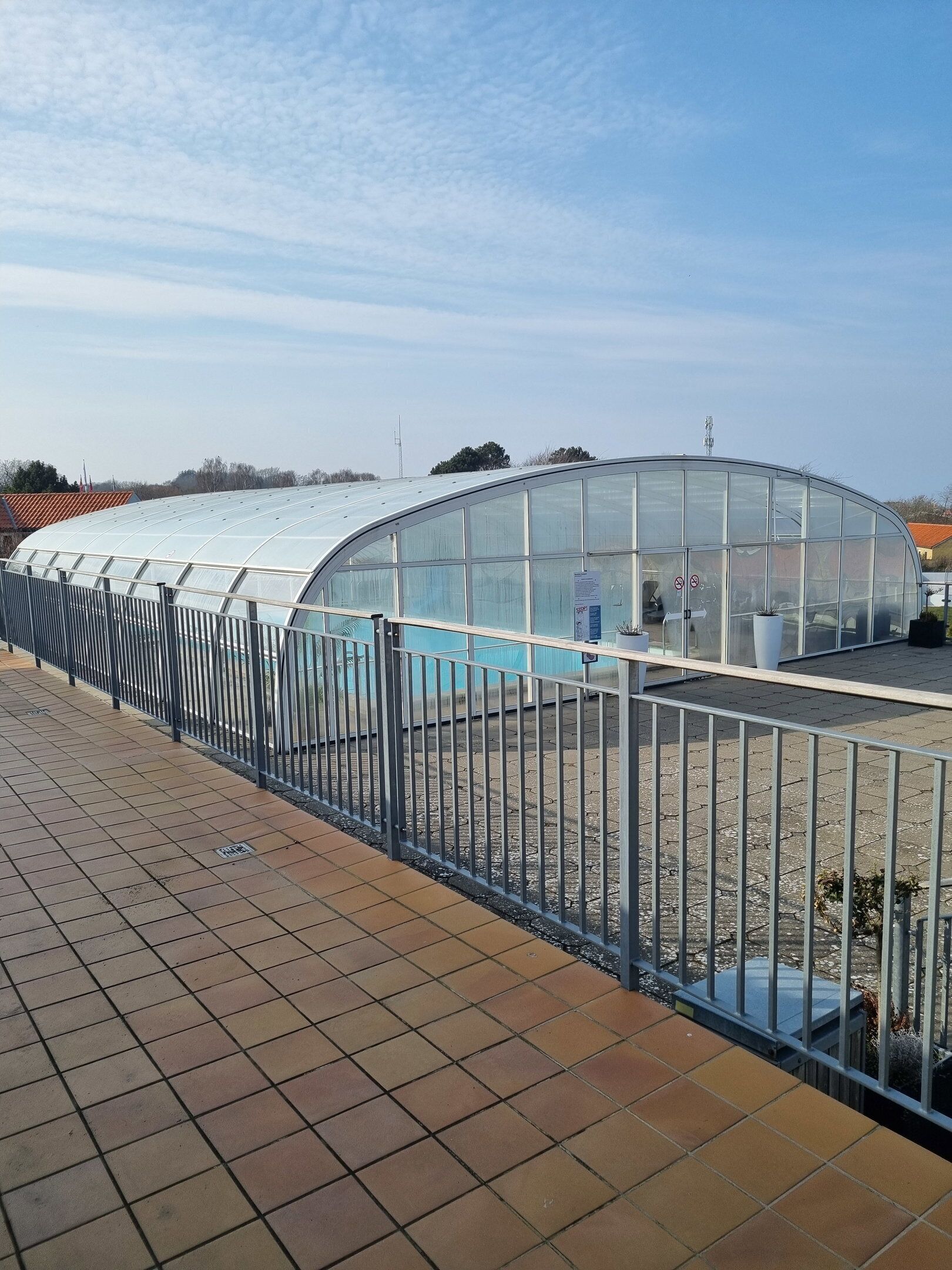 A greenhouse with a curved glass roof, surrounded by a metal fence, and a tiled walkway in the foreground.