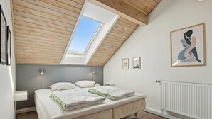 A bedroom with a sloped ceiling, a skylight, a bed with a white mattress and green blanket, two bedside lamps, and a framed abstract painting on the wall. - Summer house on beautiful Rømø! (Rømø)