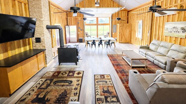 Een ruime woonkamer met een open haard, een aan een houten muur bevestigde televisie en een groot raam met uitzicht op de omgeving.