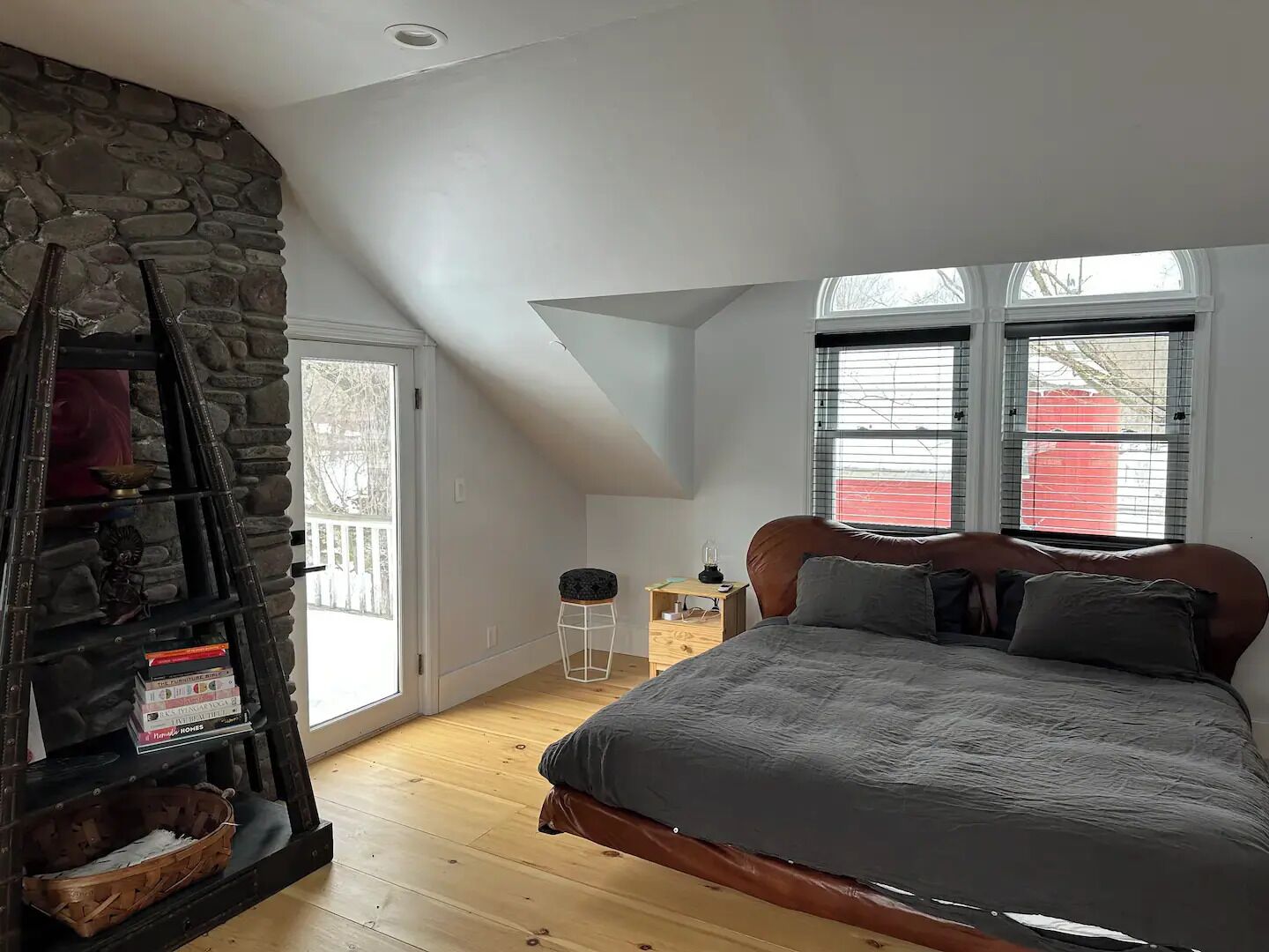 A bedroom with a stone wall, a lofted ceiling, a bed with a gray blanket, and a nightstand with a lamp.