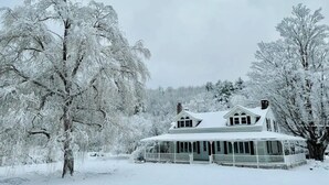 Ett snötäckt hus med en veranda och ett stort träd i förgrunden.