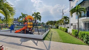 Un parque infantil con tobogán y columpios, rodeado por una cerca y palmeras.