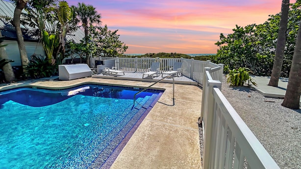 A swimming pool with lounge chairs and a deck, surrounded by palm trees and a view of the ocean at sunset. - 50 Steps to Beach, Family Friendly, Heated Pool (Indian Rocks Beach)