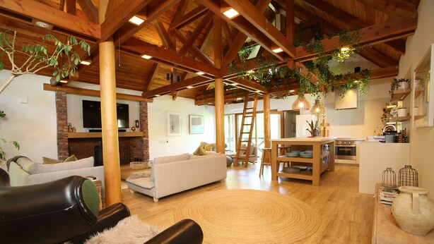 Een ruime woonkamer met een hoge plafond, houten balken, een open haard, banken, en een eethoek met een tafel en stoelen.
