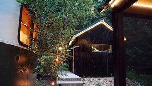 Una cabaña de madera con terraza y un pequeño porche, rodeada de árboles y un jardín.